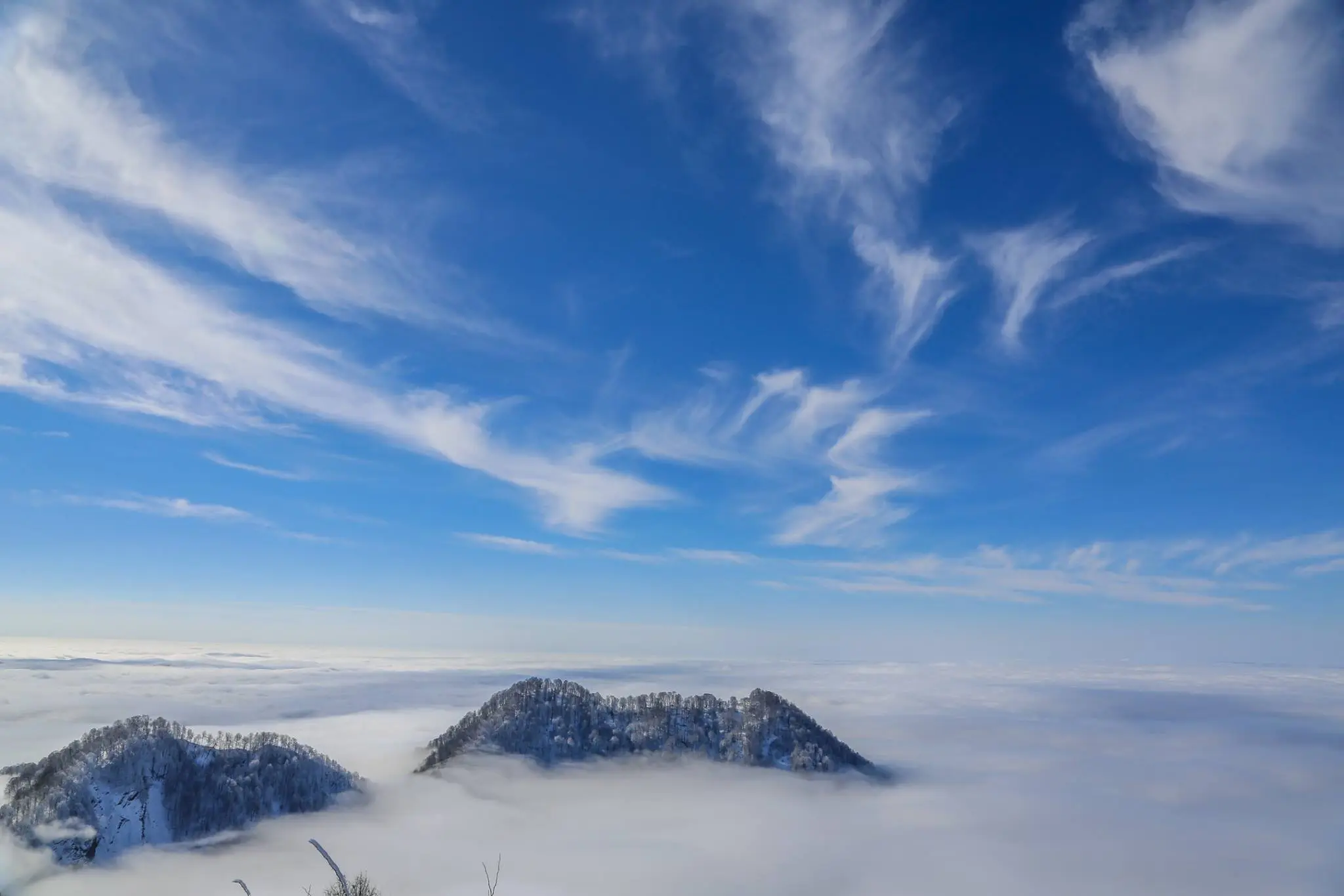A breathtaking winter view from the Gabala Cable Car shows snow-covered mountain peaks rising above a blanket of clouds under a clear blue sky.