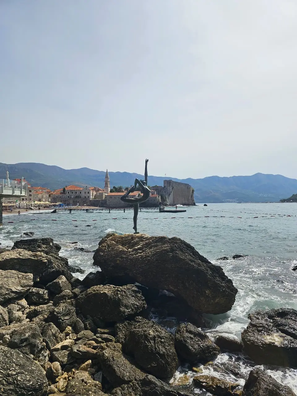Ballerina statue on Budva coast with Old Town and mountains in the background.