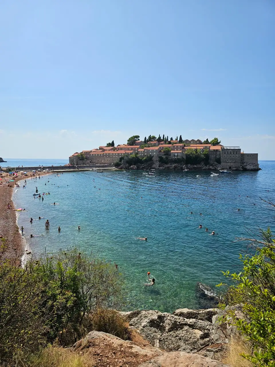 Sveti Stefan island with sandy beach and swimmers in turquoise waters.
